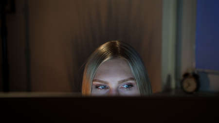 Crop view of beautiful female sitting at night at computer and looking at camera over top of monitor with dark window on blurred backgroundの写真素材