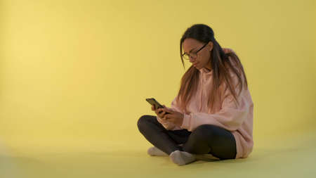 Cheerful black woman writing message on smartphone sitting on the floor in room with yellow background.の写真素材