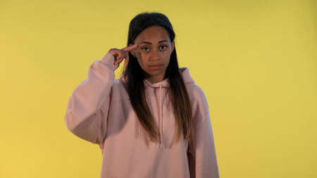 Attractive black woman twisting her index finger at temple on yellow background. She looking at camera with disagreement.の写真素材