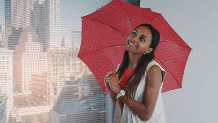 Close-up of smiling black model posing with red umbrella on bar high chair for fashion magazine photo shoot. There are skyscrapers in the background.の写真素材