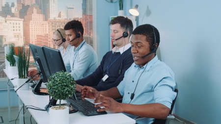 Medium long shot of Black customer support agent working in busy call center by talking to the international client. His multiethnic team also taking calls.の写真素材
