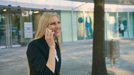 Adult blond businesswoman in suit sitting on bench and waving with hand greeting colleague on streetの写真素材