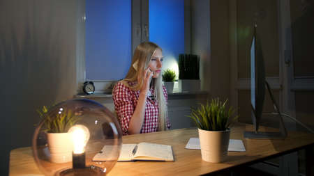 Sitting in dark room lit by small lamp at wooden desk with notebook, young blond woman in checkered shirt talking on mobile phone and looking stunned at computer monitor.の写真素材