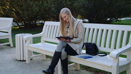 Young Beautiful Woman Sits On The Bench In Autumn Park And Works On The Tablet.の写真素材