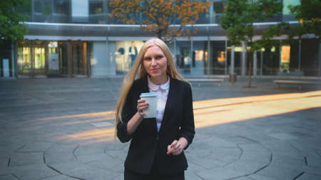 Elegant blond woman in suit and with long hair drinking coffee from white cup in patio with trees.の写真素材