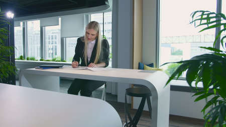 Modern blond woman in trendy suit sitting at table in light contemporary office and writing on papersの写真素材