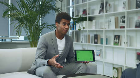 Young African-American man in suit sitting in office on sofa with anonymous colleague and making presentation with tablet with tablet with green chromakey screen.の写真素材