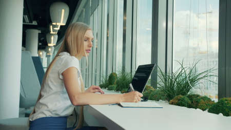 Side view of businesswoman writing on paper while sitting with laptop at table near window in modern officeの写真素材