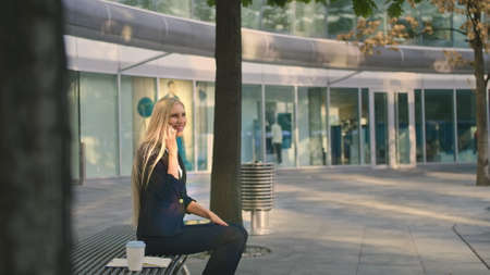 Elegant modern woman sitting on bench and waving with hand to friend while speaking on smartphone outdoorsの写真素材