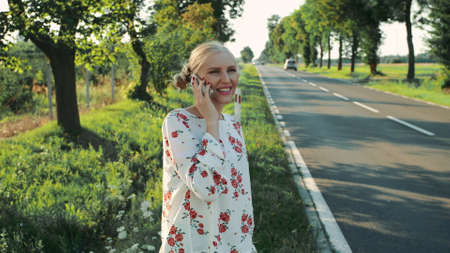 Young lady speaking on phone while hitchhiking. Side view of lovely young female talking on smartphone and waiting for car while standing on roadside during trip through countryside.の写真素材