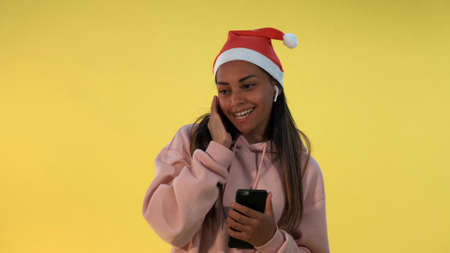 Happy african woman in Santa hat and wireless headphones listening to the music on yellow background. She singing some Christmas songs.の写真素材