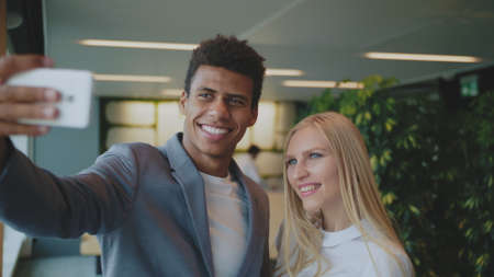 Cheerful black man with laughing blond woman taking selfie with smartphone in modern office having fun.の写真素材