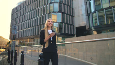 Businesswoman using phone and walking on paved street of contemporary city with new buildings of offices in sunlightの写真素材