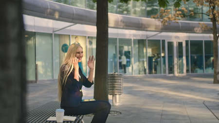 Elegant modern woman sitting on bench and waving with hand to friend while speaking on smartphone outdoorsの写真素材