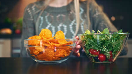 Close-up shot of lady preferring crisp to salad. Attractive young woman choosing to eat healthy crisp for lunch while sitting at table in stylish kitchen and she eats these chips.の写真素材