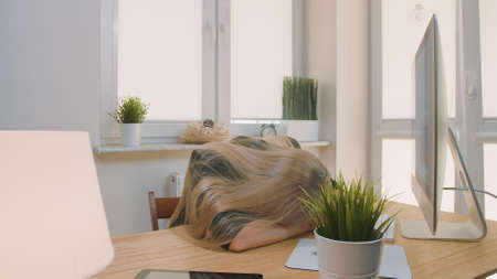 Tired blond female office worker in elegant suit relaxing lying on arms, on desk then getting up yawning and starting working on computer in light room with plants.の写真素材