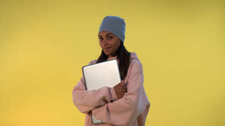 Portrait of African girl in cap hugging her computer laptop and smiling on yellow background.の写真素材