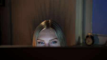 Crop view of beautiful female sitting at night at computer and looking at camera over top of monitor with dark window on blurred backgroundの写真素材