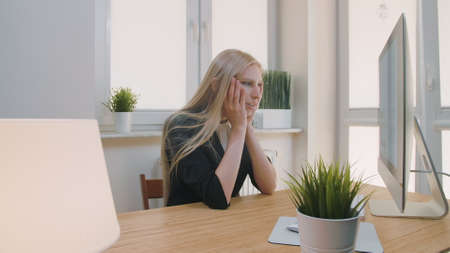 Blond sad female in elegant clothes clutching head after failure or bad news sitting in light office at desk with computerの写真素材