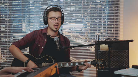 Musician in headphone and trendy casual clothes recording his new song in home studio. He also playing guitar while his friend accompanying him.の写真素材