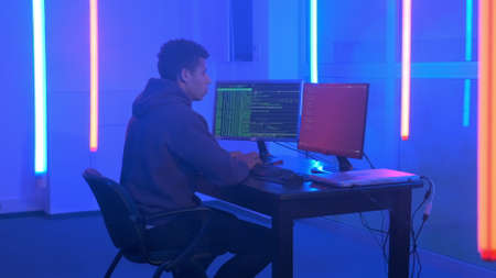 Young african man writing hacker programs in dark room with neon lights. He working on computer with two monitors.の写真素材