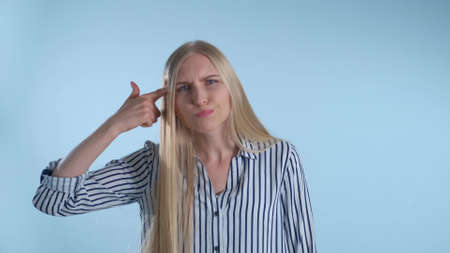 Upset young woman beating yourself on forehead and shooting herself in the head with finger gun gesture. There is a blue background.の写真素材