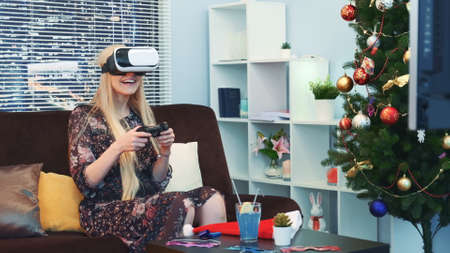 Medium close up of nice girl playing a game in virtual reality glasses in front of TV on Christmas. There are skyscrapers in the background.Future technology concept.の写真素材