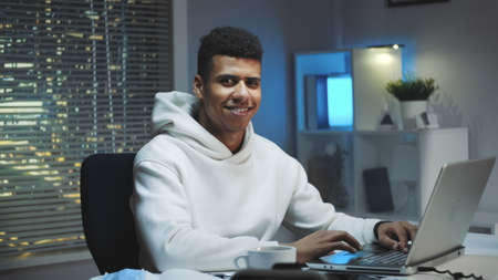 Handsome black man smiling to the camera while working home on computer. Skyscrapers in the...