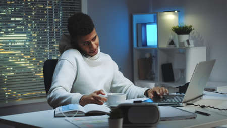 Mixed-race man working on computer, speaking by smartphone and drinking a cup of tea. Home office with skyscrapers in the background.の写真素材