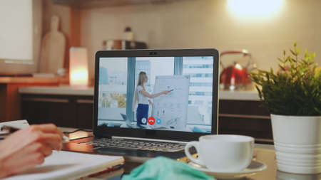 Online video conference on laptop with female speaker in home office. In the foreground: writing hands, medical mask and a cup.の写真素材