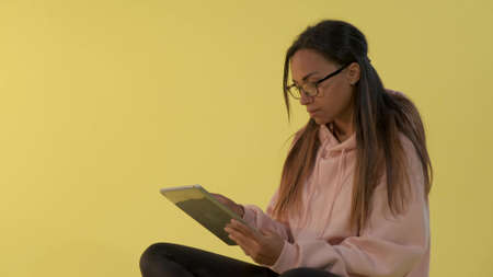 Black woman in eyeglasses working on tablet on yellow background. She sitting on the floor.の写真素材