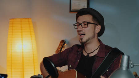 Close up of bearded guy in hat emotionally singing into microphone and playing guitar in home studio. He is rehearsing in the evening.の写真素材