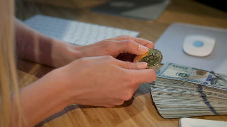 Crop view of hands of woman holding shiny metal bitcoin sitting at wooden desk with wad of cash nearby at nightの写真素材