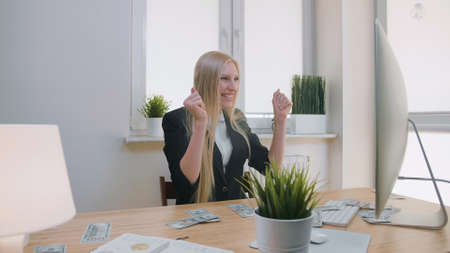 Elegant blond female sitting at desk at workplace with computer holding in hands bundle of cash looking astonished at monitor, and throwing money in air and happily making yes gesture.の写真素材