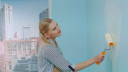Portrait shot of a young beautiful woman painting a wall at office. She uses white paint and work with a roller. Office renovations.の写真素材