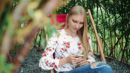 Young woman using smartphone in plant gazebo. Beautiful young lady smiling and typing message on smartphone while sitting inside amazing plant gazebo in park.の写真素材