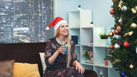 Beautiful girl in Santa hat is waiting and emotionally celebrating the New Years coming with a glass of drink in her hands, blowing in party whistle. There are skyscrapers in the background.の写真素材
