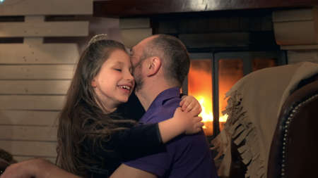 Happy father and small daughter embracing each other on armchair near the fireplace. They love to spend free time in the evening together.の写真素材
