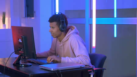 Persistent black gamer playing online shooter on pc in neon room. He spending his leisure time at home.の写真素材