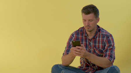 Cheerful man in checkered shirt sitting on the floor and chatting on smartphone. There is yellow background.の写真素材