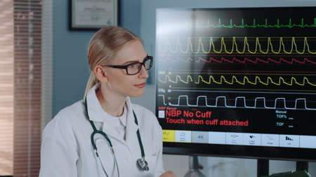 Close-up of beautiful female doctor in lab coat discussing EKG data with colleague showing on big...