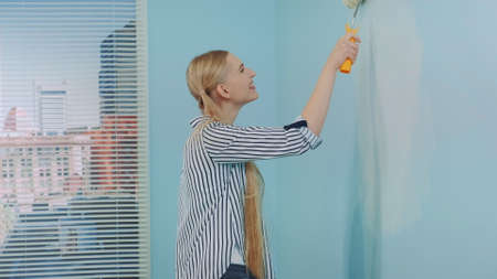 Woman makes repairs in the apartment dancing and smiling. She uses white paint and work with a roller. In the background there are skyscrapers at day.の写真素材