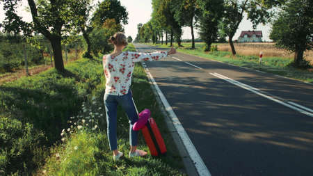 Young woman hitchhiking on countryside road. Back view of young female with red suitcase standing on roadside and hitchhiking while travelling through countryside.の写真素材