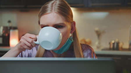 Close-up of business woman working at the computer and shifting medical mask from face todrink something. She working hard on her work project at home.の写真素材