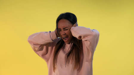 Desperate african woman screaming and covering ears on yellow background. She is annoyed with loud noise.の写真素材