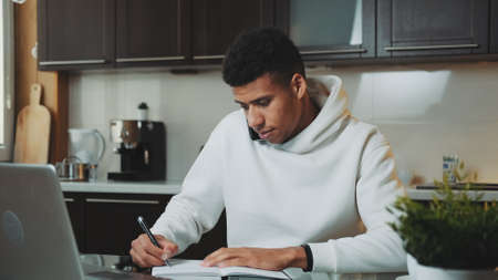 Mixed-race man speaking by smartphone and working on computer at home kitchen. He is dressed in white hoodie.の写真素材