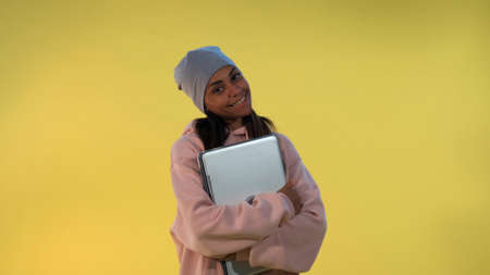 Portrait of African girl in cap hugging her computer laptop and smiling on yellow background.の写真素材