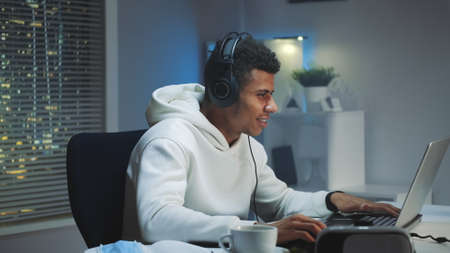 Black gamer in white hoodie and with headphones playing games on computer in the evening. He is passionate and smiled.の写真素材