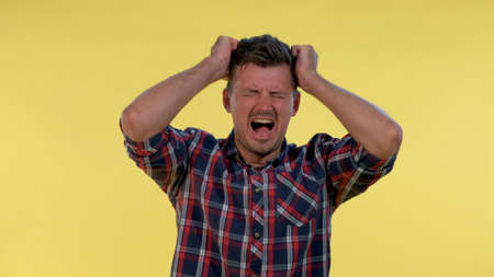 Young man in checkered shirt putting fists on head and screaming in pain. He has a terrible headache.の写真素材