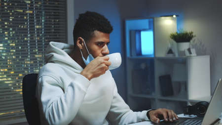 Medium shot of Handsome multiracial man drinking a cup of coffee and working on computer at night. There are skyscrapers in the background.の写真素材